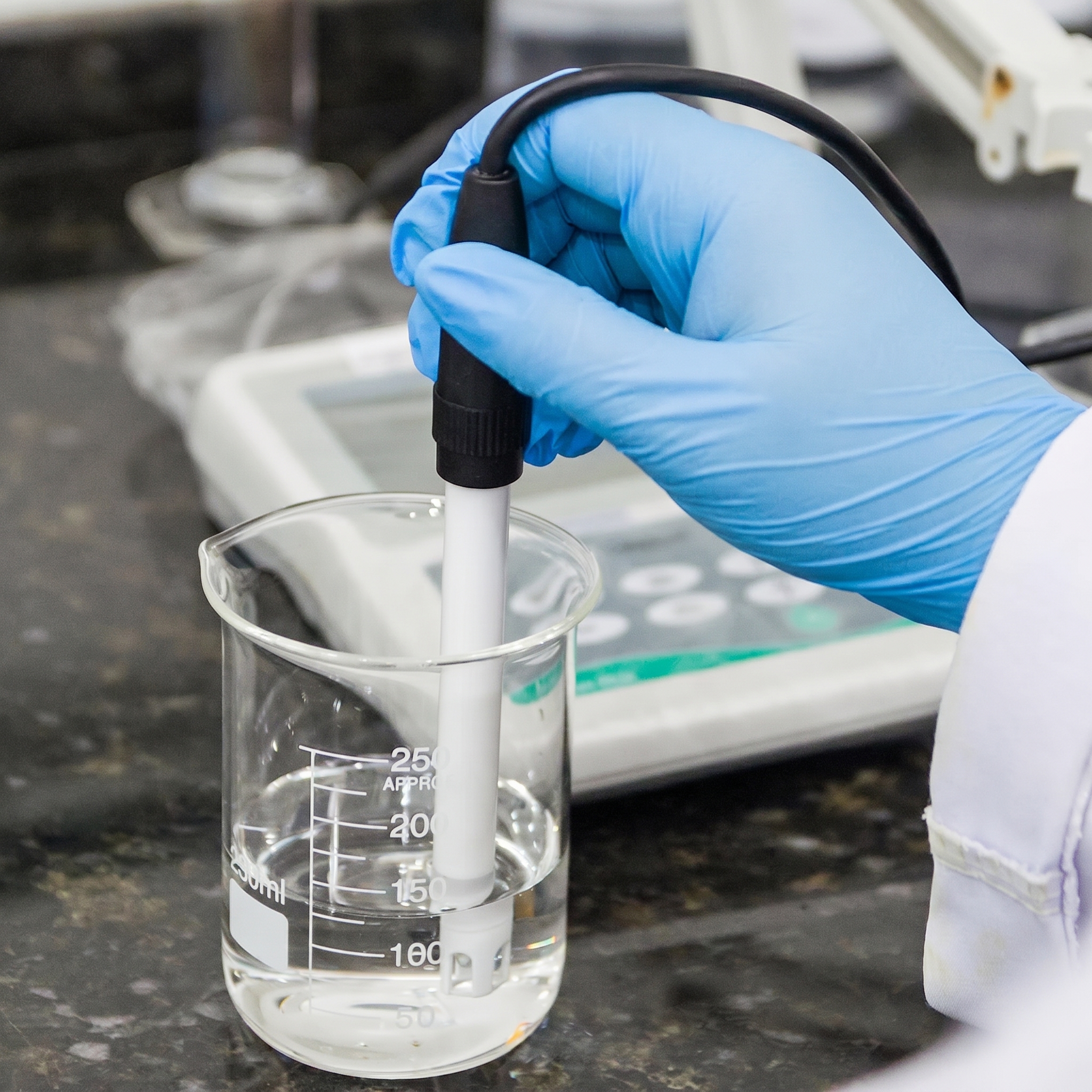 Gloved researcher measuring water quality with a probe in a beaker beside a digital meter